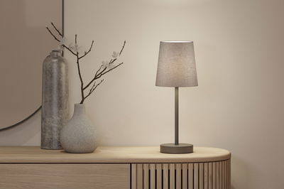 Moderne tafellamp met grijze stoffen kap en metalen voet op licht houten dressoir met decoratie.