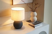 Keramik-Tischlampe mit hellem Stoffschirm und weiße Vase mit Trockenblumen auf weißem Sideboard