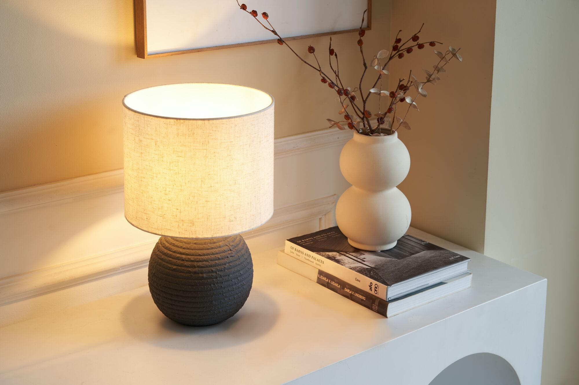 Keramik-Tischlampe mit hellem Stoffschirm und weiße Vase mit Trockenblumen auf weißem Sideboard