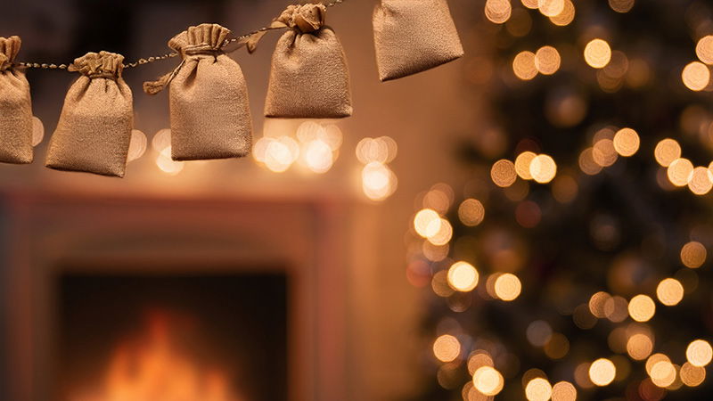 Beige Stoffbeutel an Schnur vor unscharfem Kaminfeuer und Weihnachtsbaum mit Lichterkette