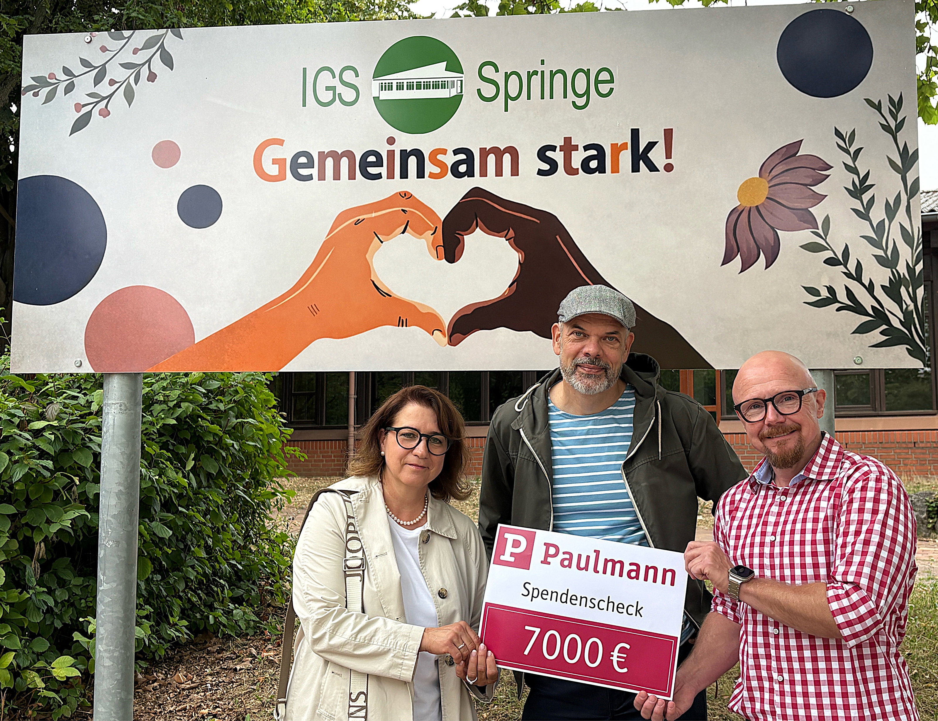 Drei Personen übergeben einen Spendenscheck über 7000 Euro vor IGS Springe Banner mit Herzsymbol