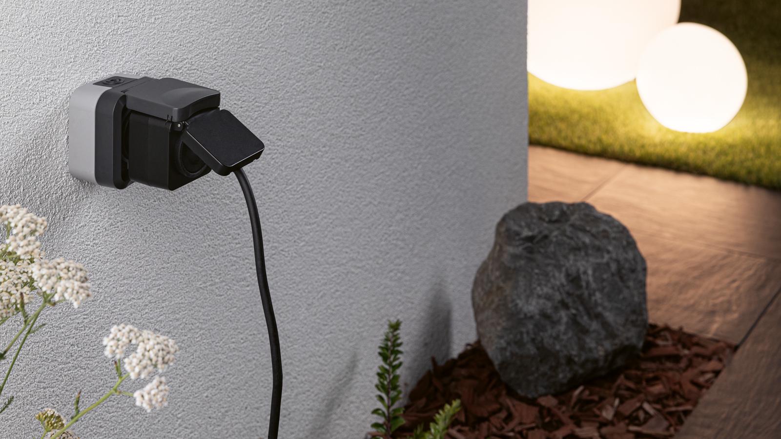 Black outdoor socket with flap cover on grey wall, garden lighting with LED spheres in background