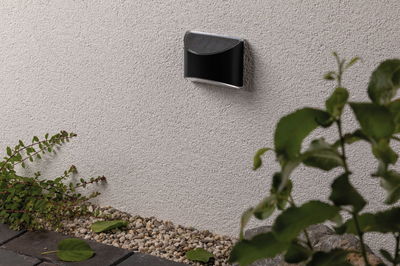 Schwarze LED-Wandleuchte aus Metall an weißer Außenwand mit Kies und Pflanzen im Garten