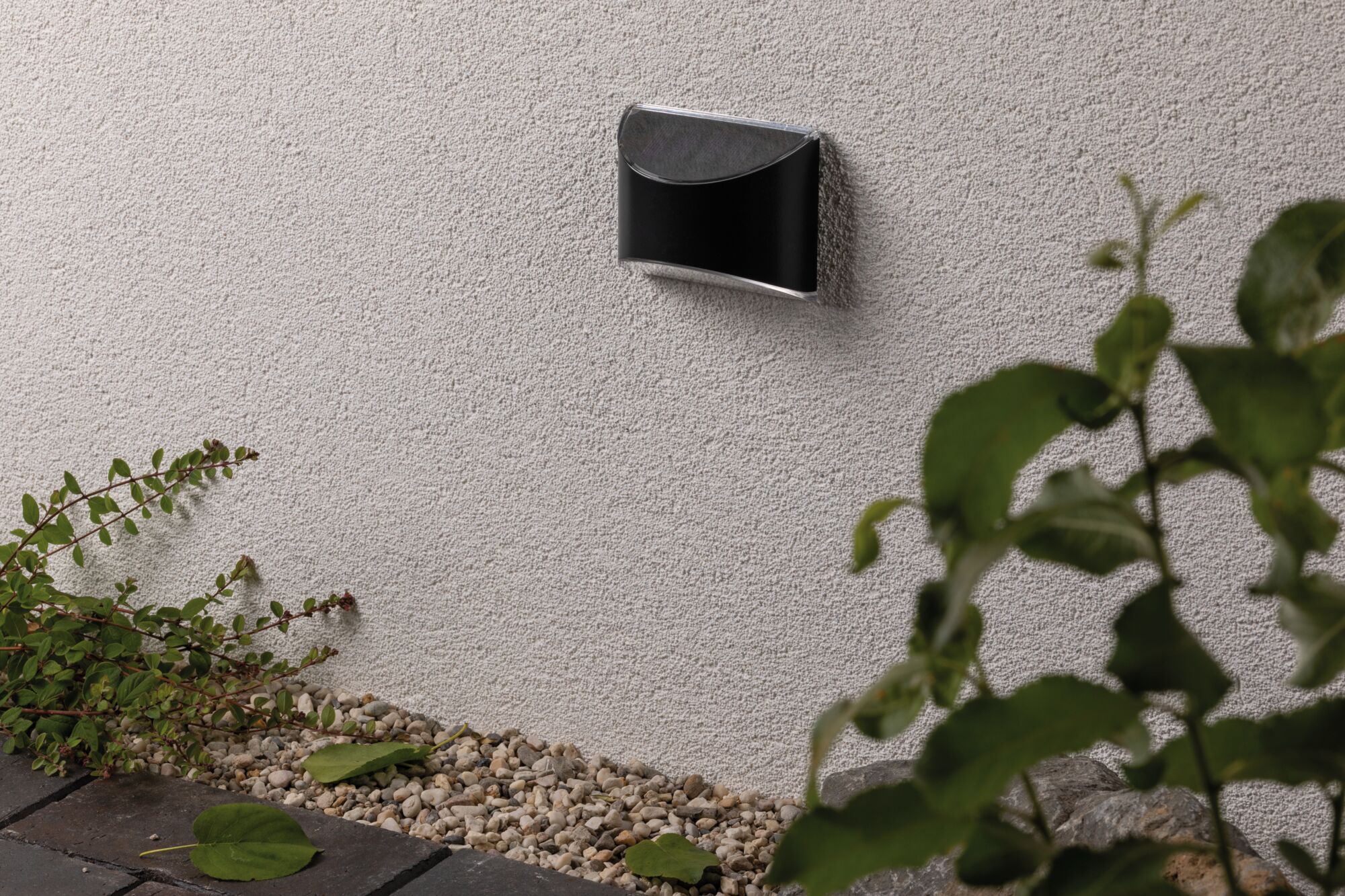 Black metal LED wall light on white exterior wall with gravel and garden plants