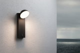 Modern black metal LED wall light with round head on white wall, outdoor lighting fixture