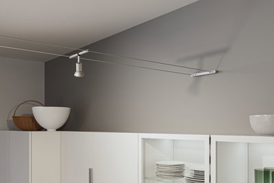 Modern white metal cable spotlight on grey wall above white kitchen cabinet with dishes.