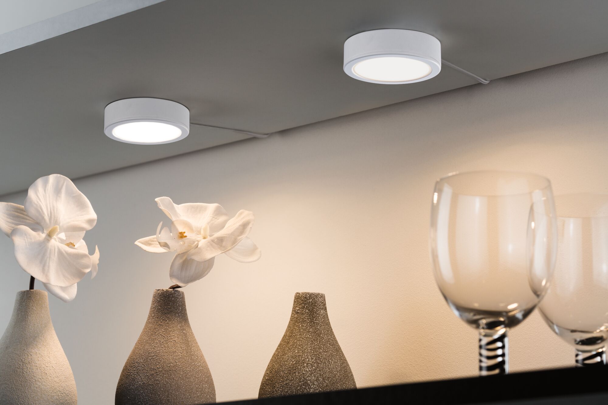 Zwei weiße runde LED-Deckenleuchten mit warmweißem Licht über Vasen und Gläsern aus Keramik und Glas