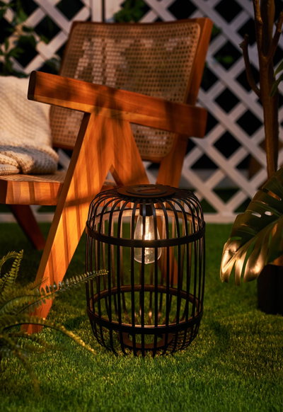 Lanterne de jardin en métal noir avec ampoule sur gazon vert près d'une chaise en bois et plantes.