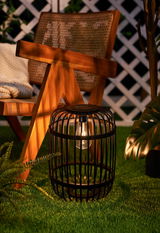 Black metal garden lantern with bulb on green grass beside wooden chair and plants.