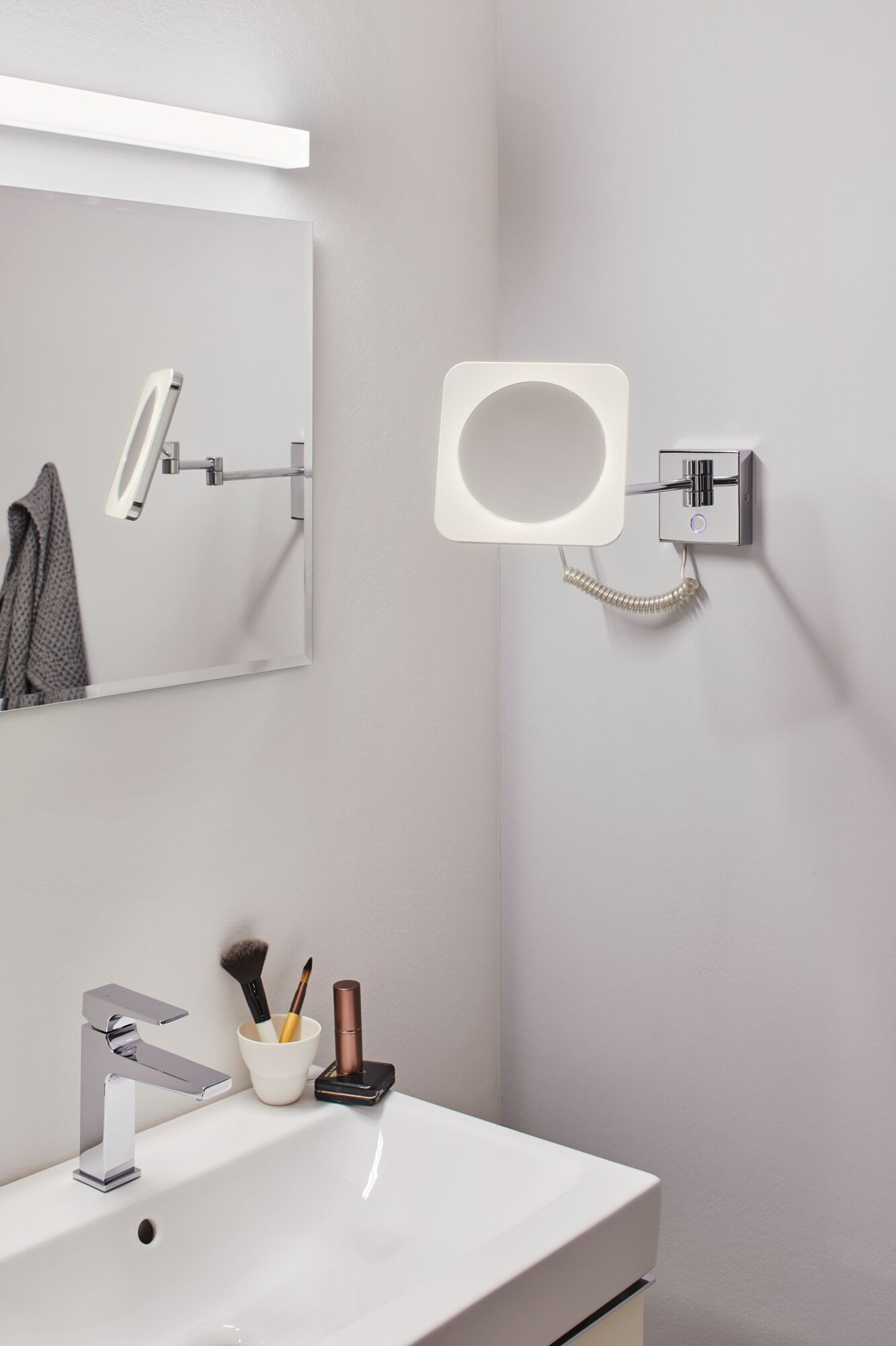 Modern LED wall mirror with chrome arm and square light ring in bathroom setting.
