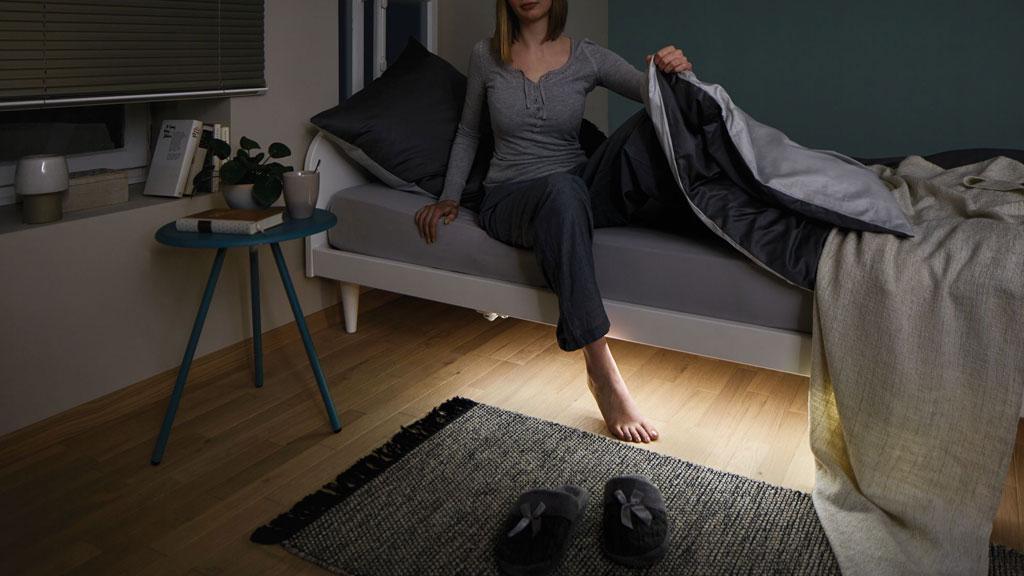 Kvinde sidder på gråt seng med indirekte LED-gulvbelysning, hyggeligt soveværelse med tæppe og natbord.