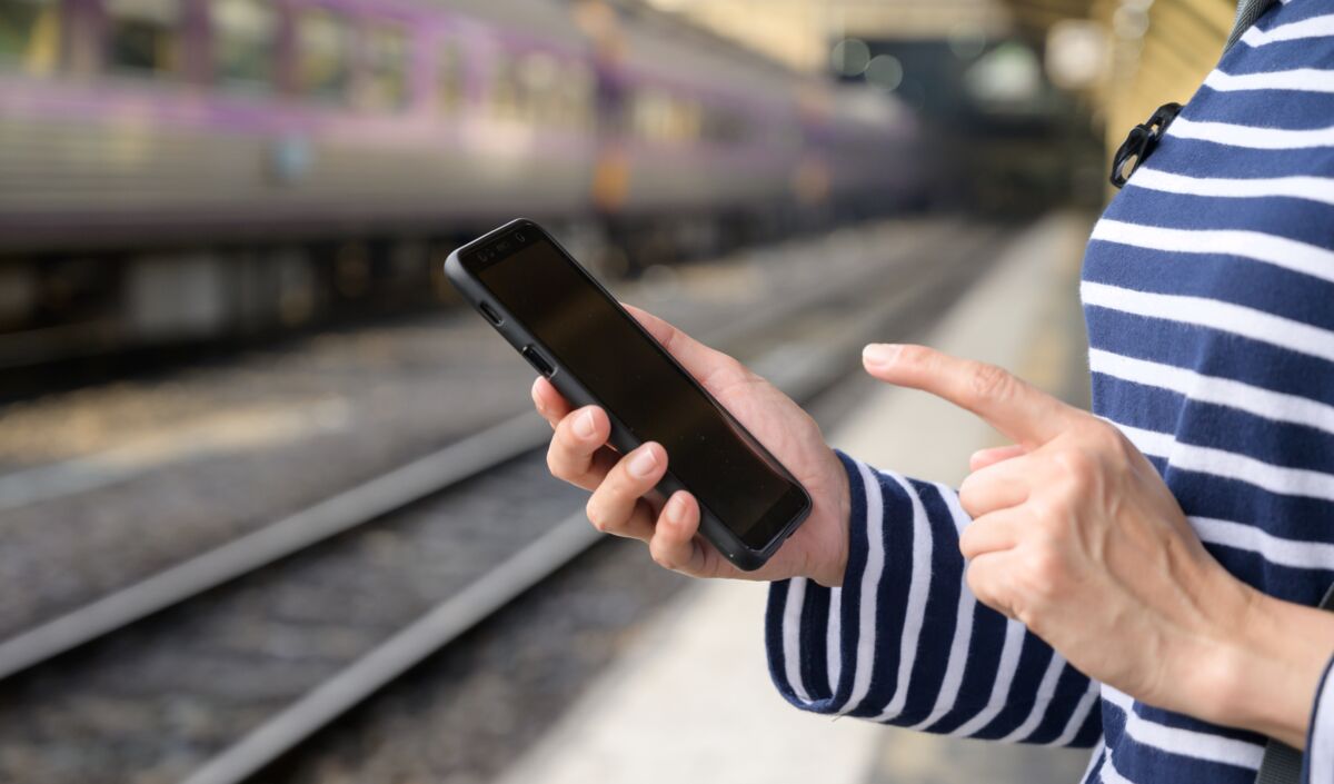 Person mit gestreiftem Pullover hält schwarzes Smartphone am Bahnsteig neben Gleisen