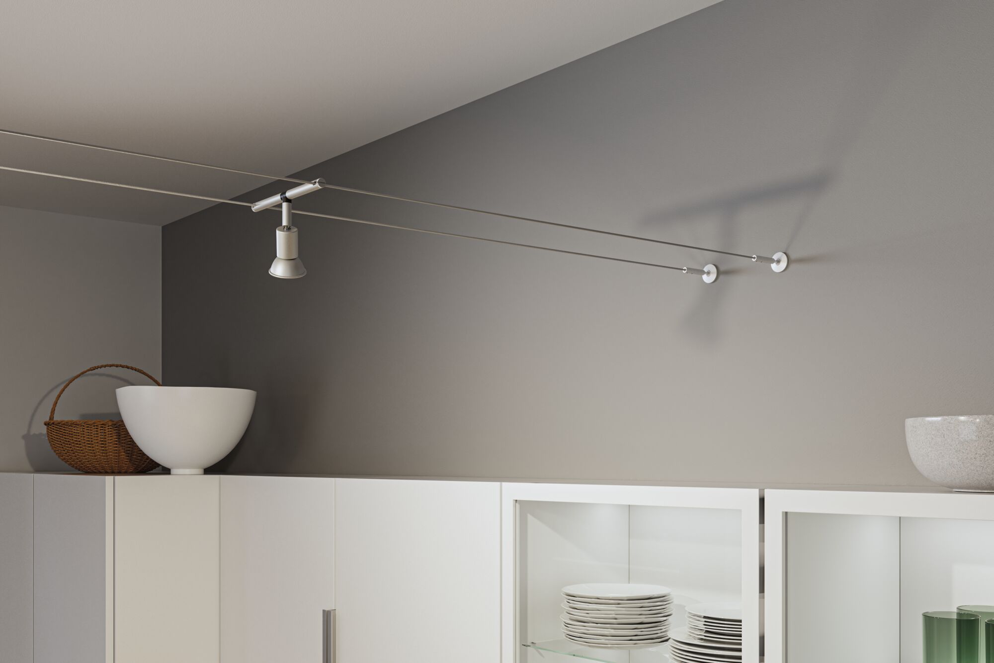 Modern white metal cable spotlight on grey wall above white kitchen cabinet with dishes.