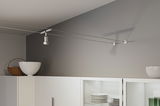 Modern white metal cable spotlight on grey wall above white kitchen cabinet with dishes.