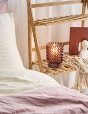 Pink glass candle holder with zigzag pattern on bamboo shelf beside bed with pink bedding