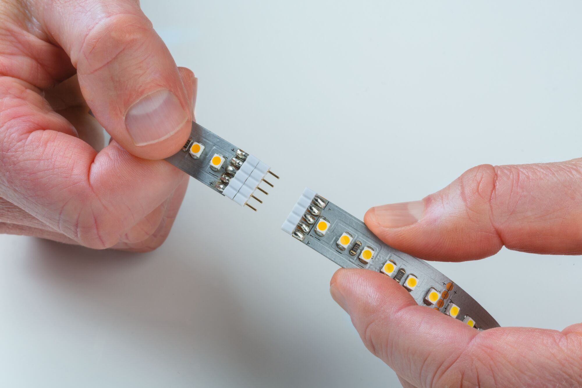 Deux mains connectant une bande LED avec LEDs jaunes et contacts métalliques sur fond blanc