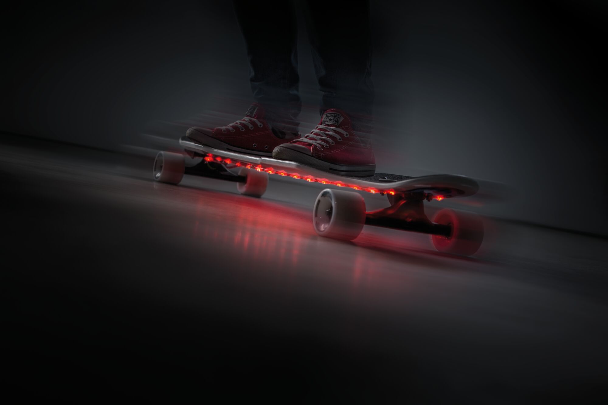 Skateboard with red LED strips under deck, white wheels, person wearing red shoes riding indoors.