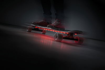 Skateboard with red LED strips under deck, white wheels, person wearing red shoes riding indoors.