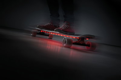 Skateboard with red LED strips under deck, white wheels, person wearing red shoes riding indoors.