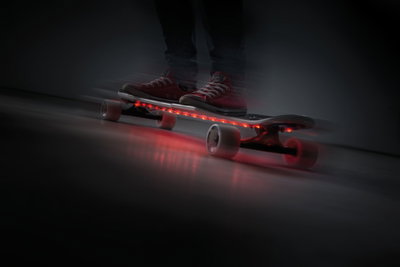 Skateboard avec bandes LED rouges sous la planche, roues blanches, personne en chaussures rouges à l'intérieur.