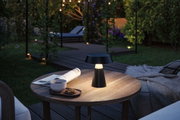 Schwarze LED-Tischlampe aus Metall auf Holztisch im Garten mit Tasse, Keks und Zeitung bei Abendlicht