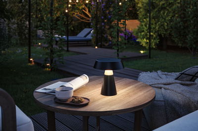 Schwarze LED-Tischlampe aus Metall auf Holztisch im Garten mit Tasse, Keks und Zeitung bei Abendlicht