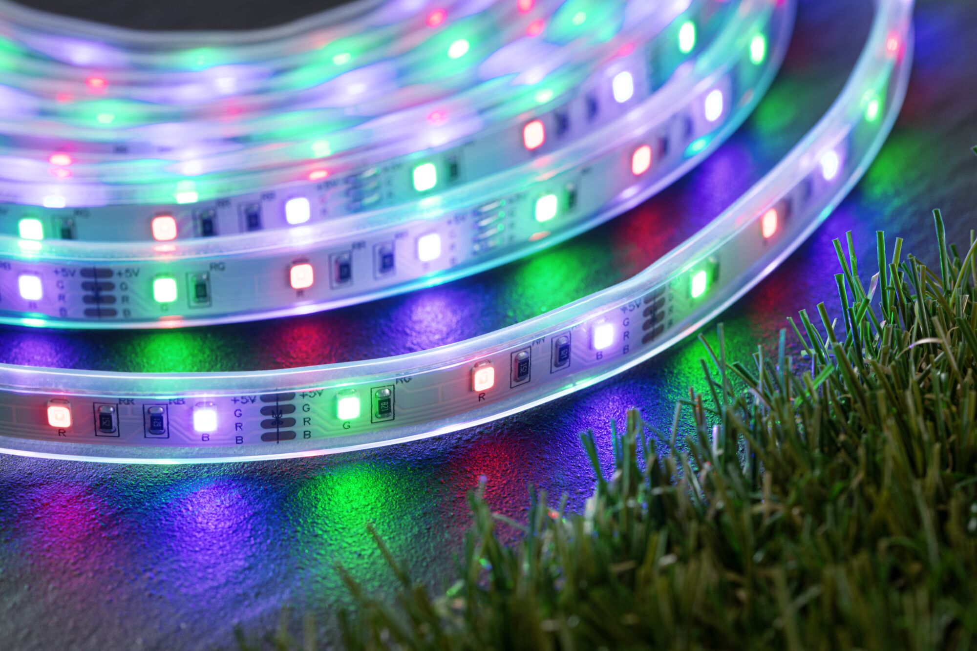 Colorful RGB LED light strips on dark surface next to green grass for decorative lighting.