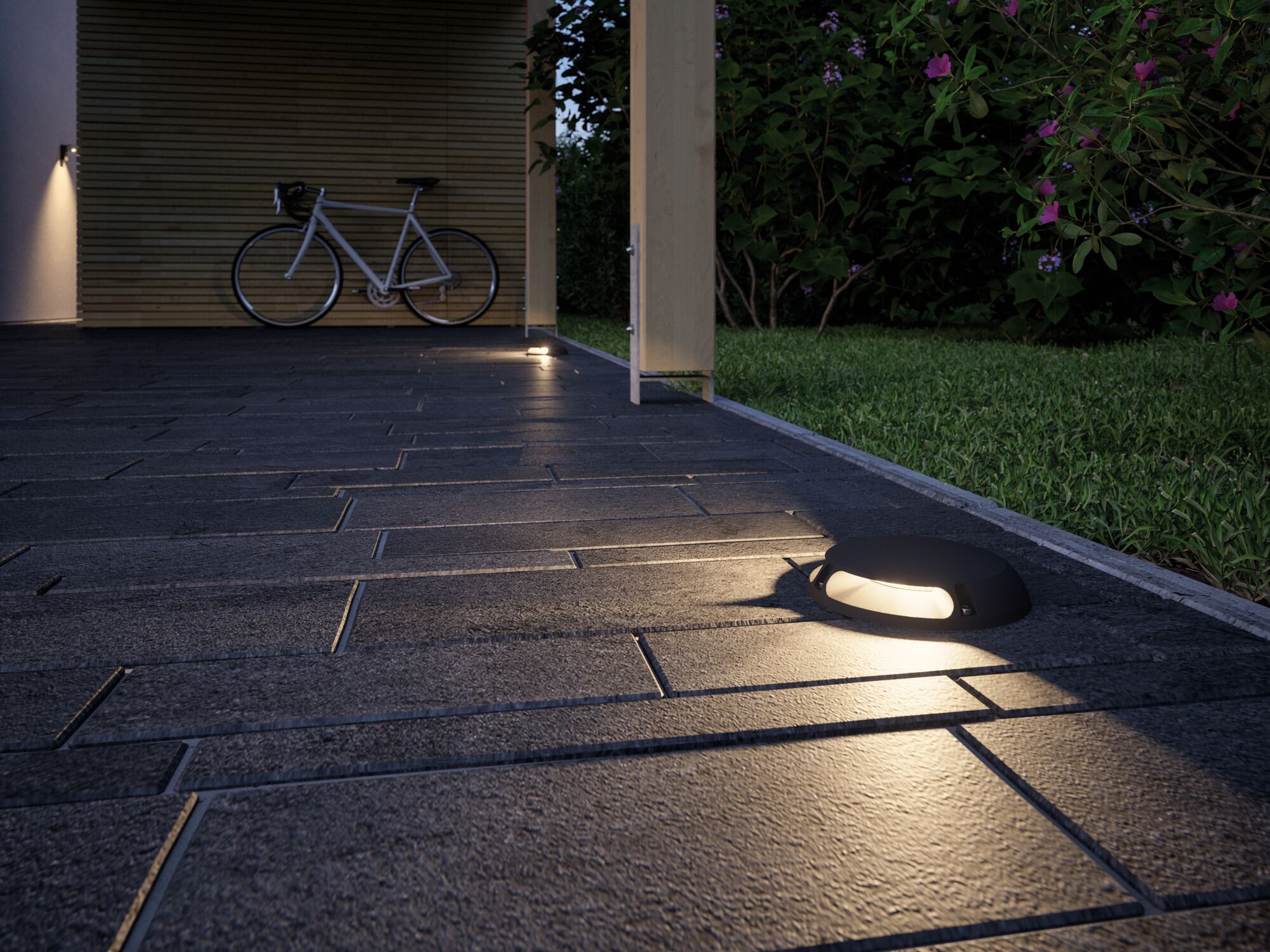 Schwarze LED-Bodenleuchte auf grauem Steinpflaster neben grünem Rasen und Fahrrad im Hintergrund
