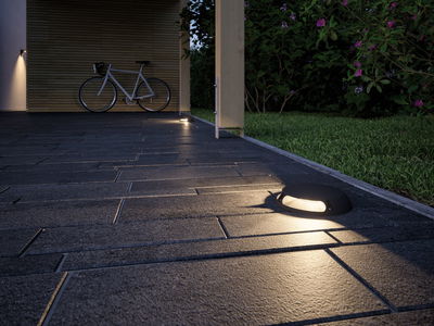 Schwarze LED-Bodenleuchte auf grauem Steinpflaster neben grünem Rasen und Fahrrad im Hintergrund