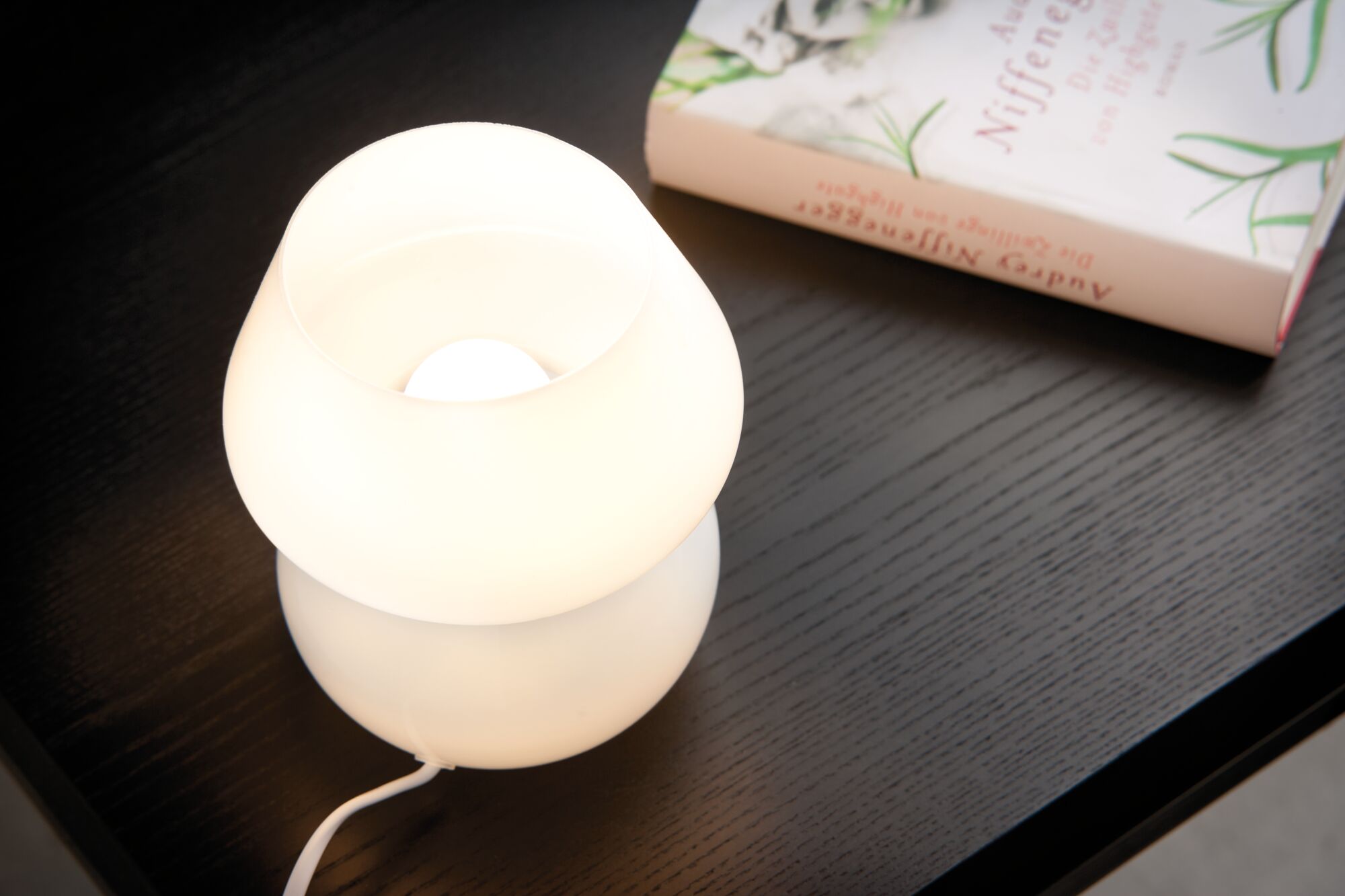White glass table lamp with warm light on black wooden table next to book for home lighting