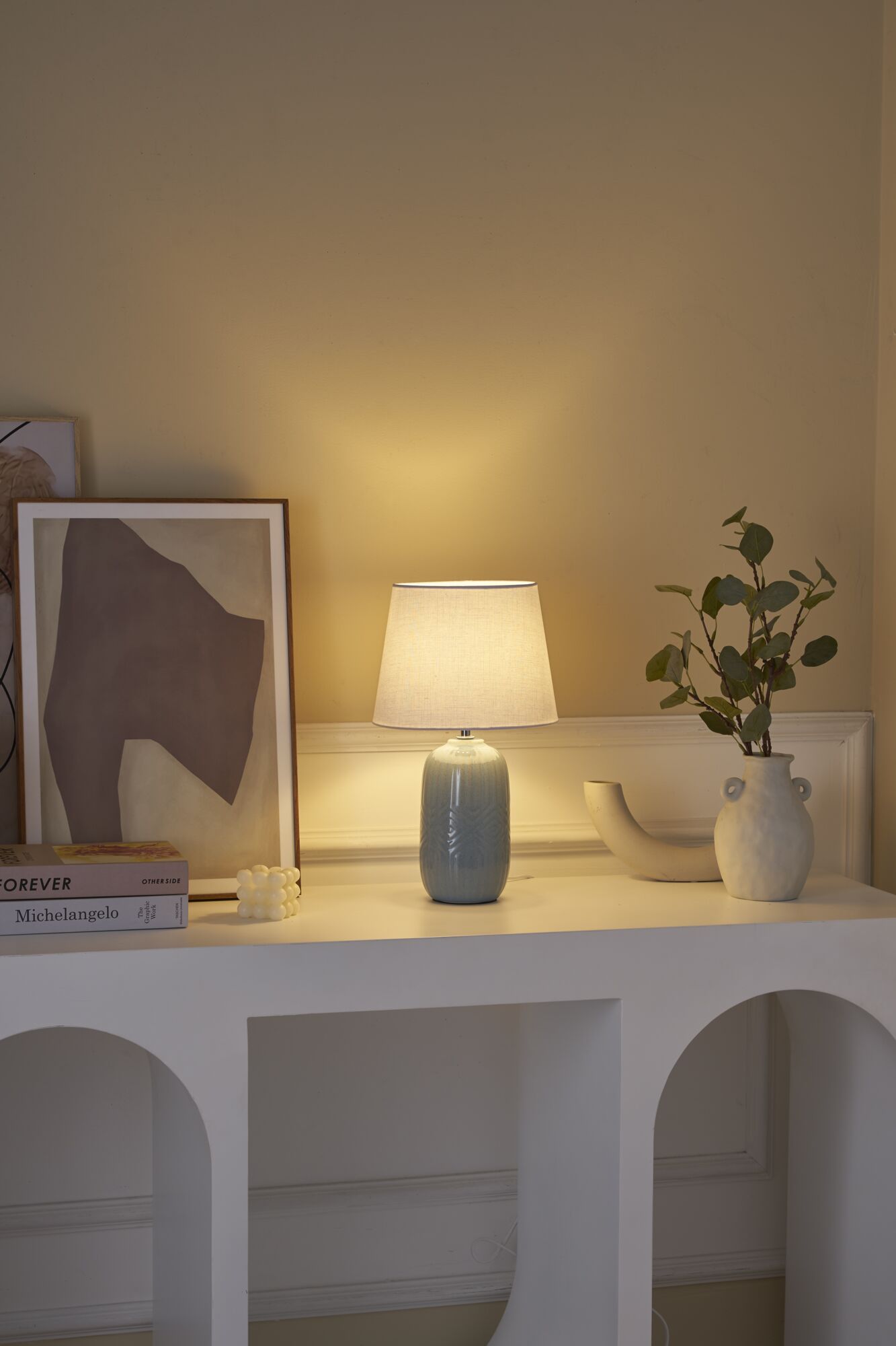 Light blue ceramic table lamp with white fabric shade on white console table with decor and books