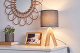 Moderne Tischlampe mit Holzfuß und grauem Stoffschirm auf weißem Sideboard mit Dekoration.