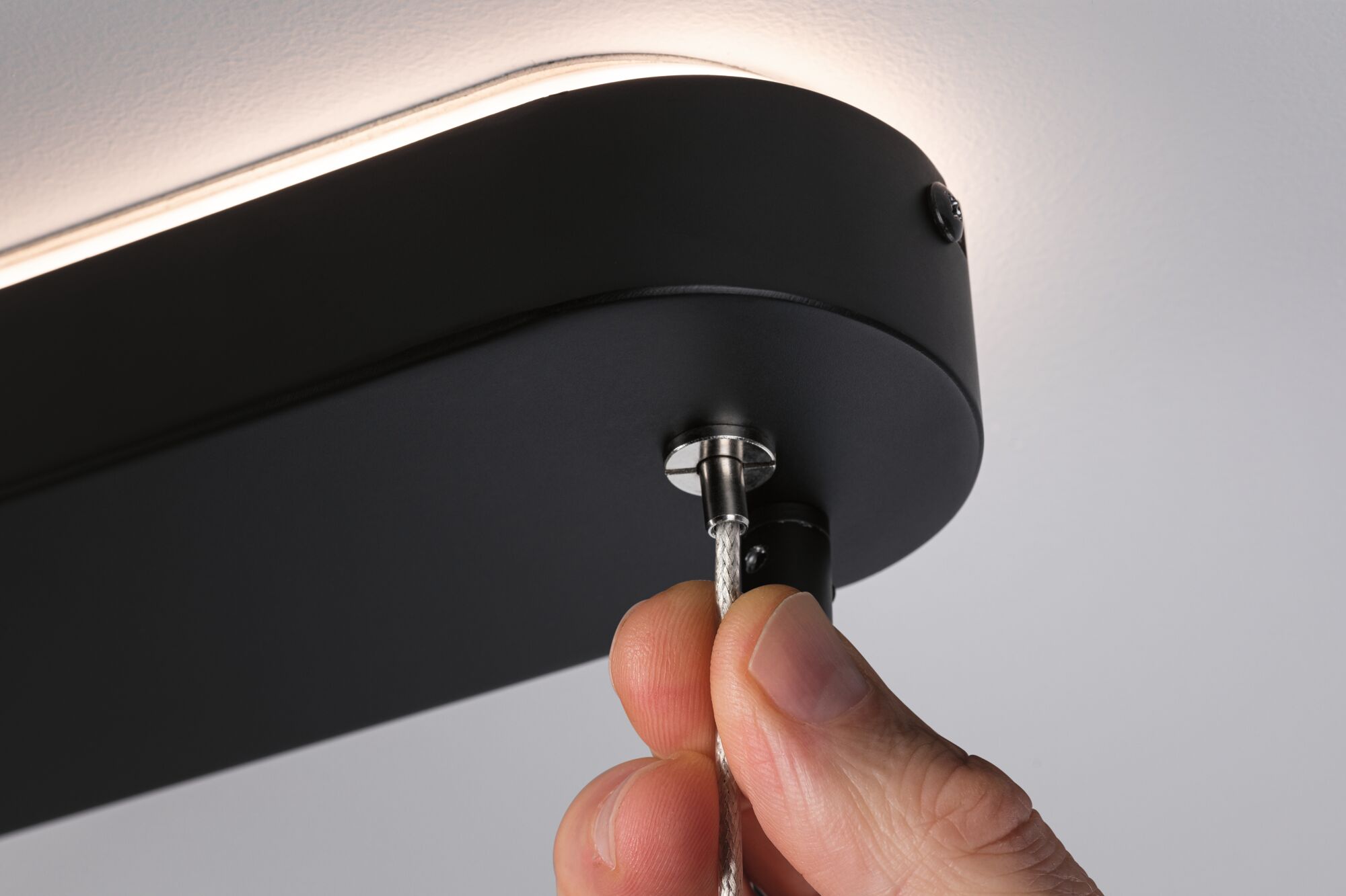 Hand attaching silver steel cable to black LED ceiling light with indirect illumination.