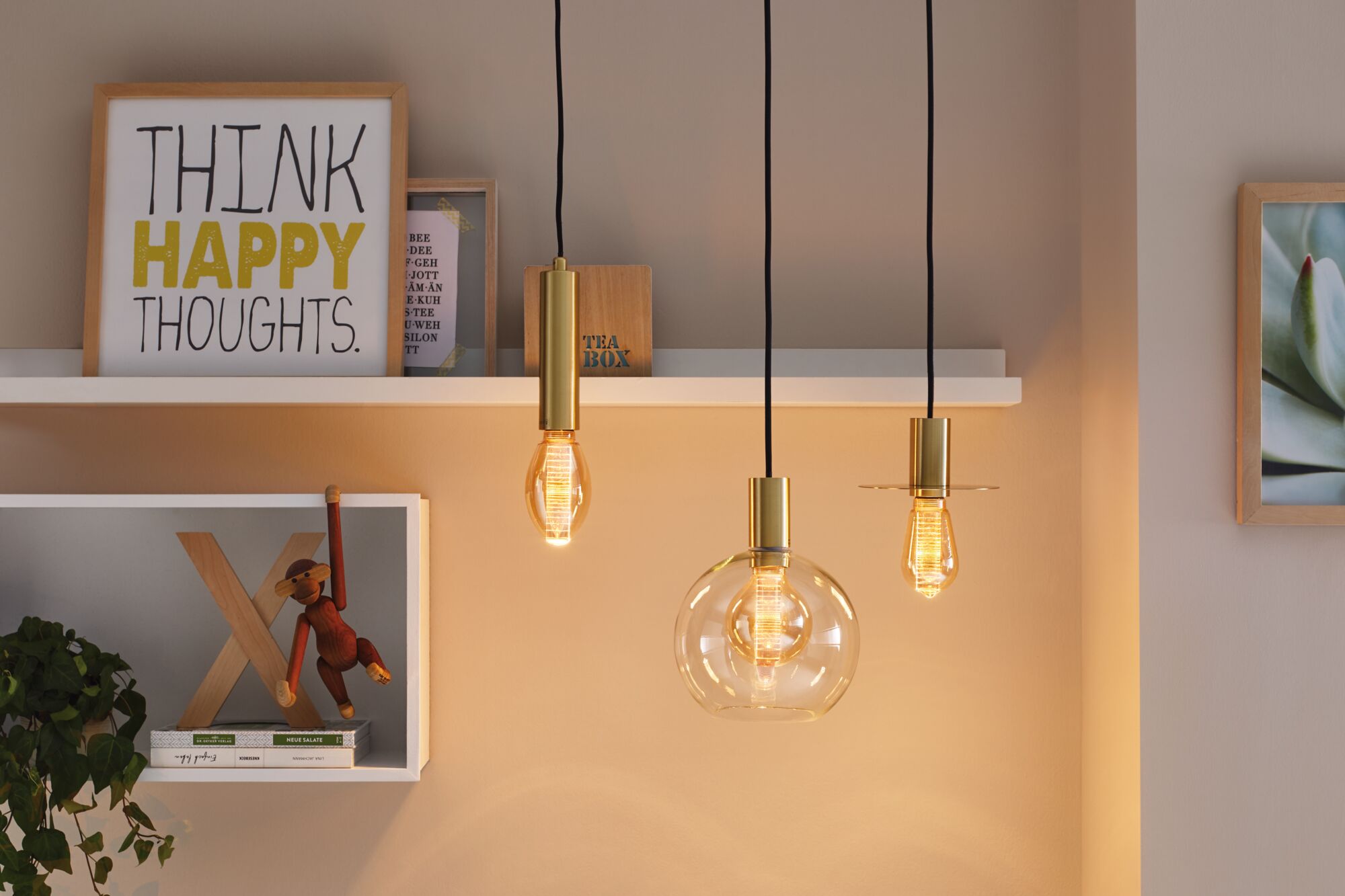 Drie gouden LED-hanglampen met warm licht tegen een beige muur en decoratieve planken.