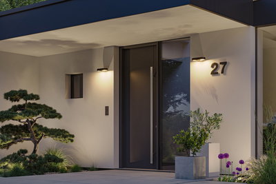 Moderne Hausfassade mit schwarzer Tür, grauem Pflanzkübel und warmweißen Außenwandleuchten.