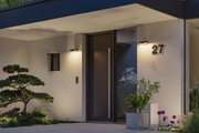 Moderne Hausfassade mit schwarzer Tür, grauem Pflanzkübel und warmweißen Außenwandleuchten.