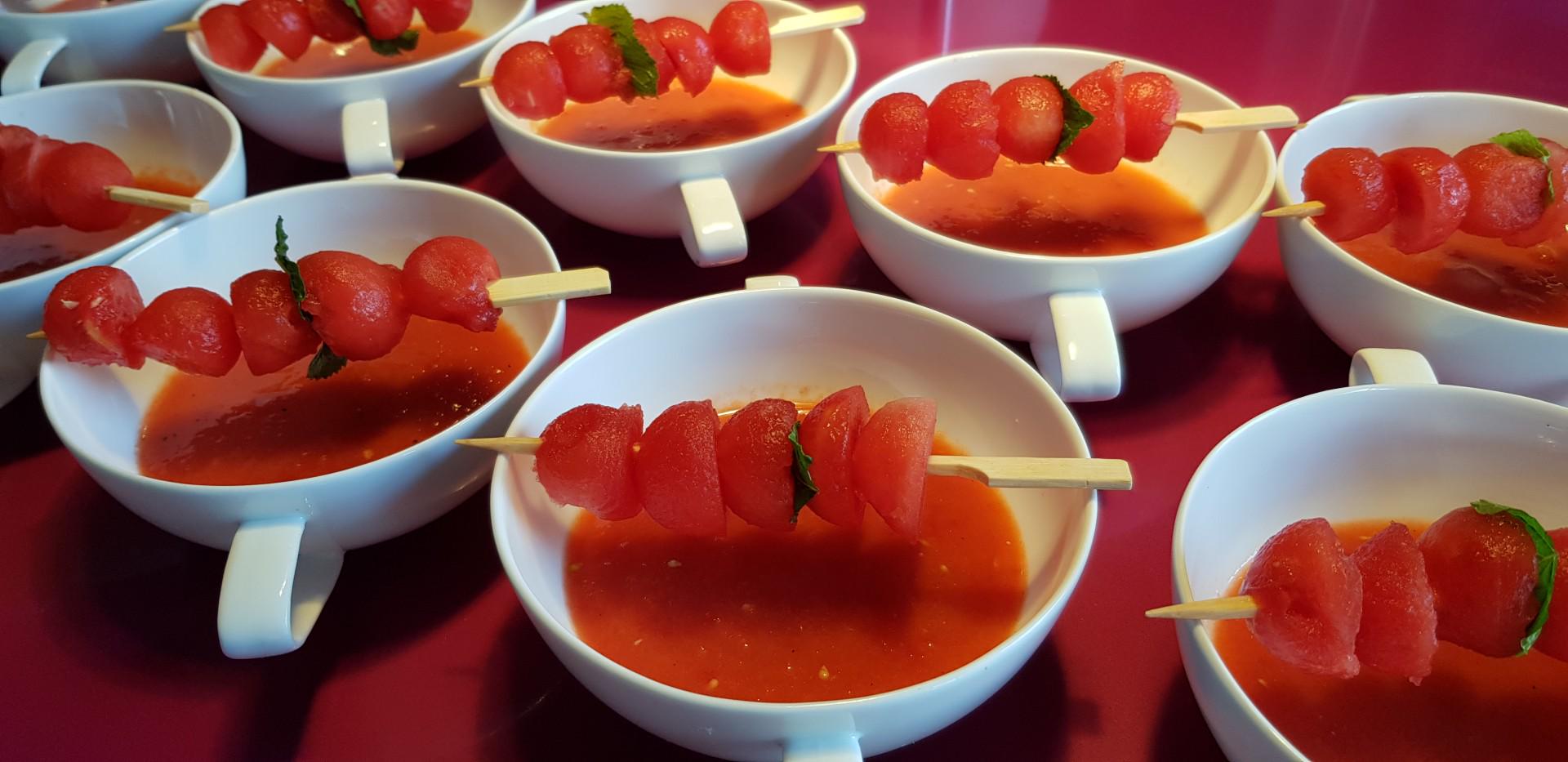Mehrere weiße Tassen mit roter Tomatensuppe und Spießen aus Wassermelonenbällchen und Minze.