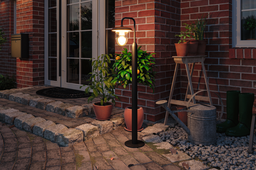 Schwarze Außen-Stehleuchte mit warmweißem Licht neben Topfpflanzen vor rotem Backsteinhaus.