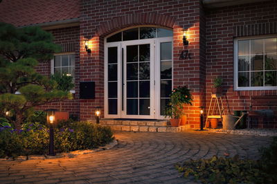 Illuminated house entrance with white doors, red brick walls, and warm outdoor lights along garden path.