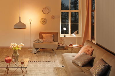 Gezellige woonkamer met beige meubels, warm wit licht, decoratieve kussens en wandklokken.