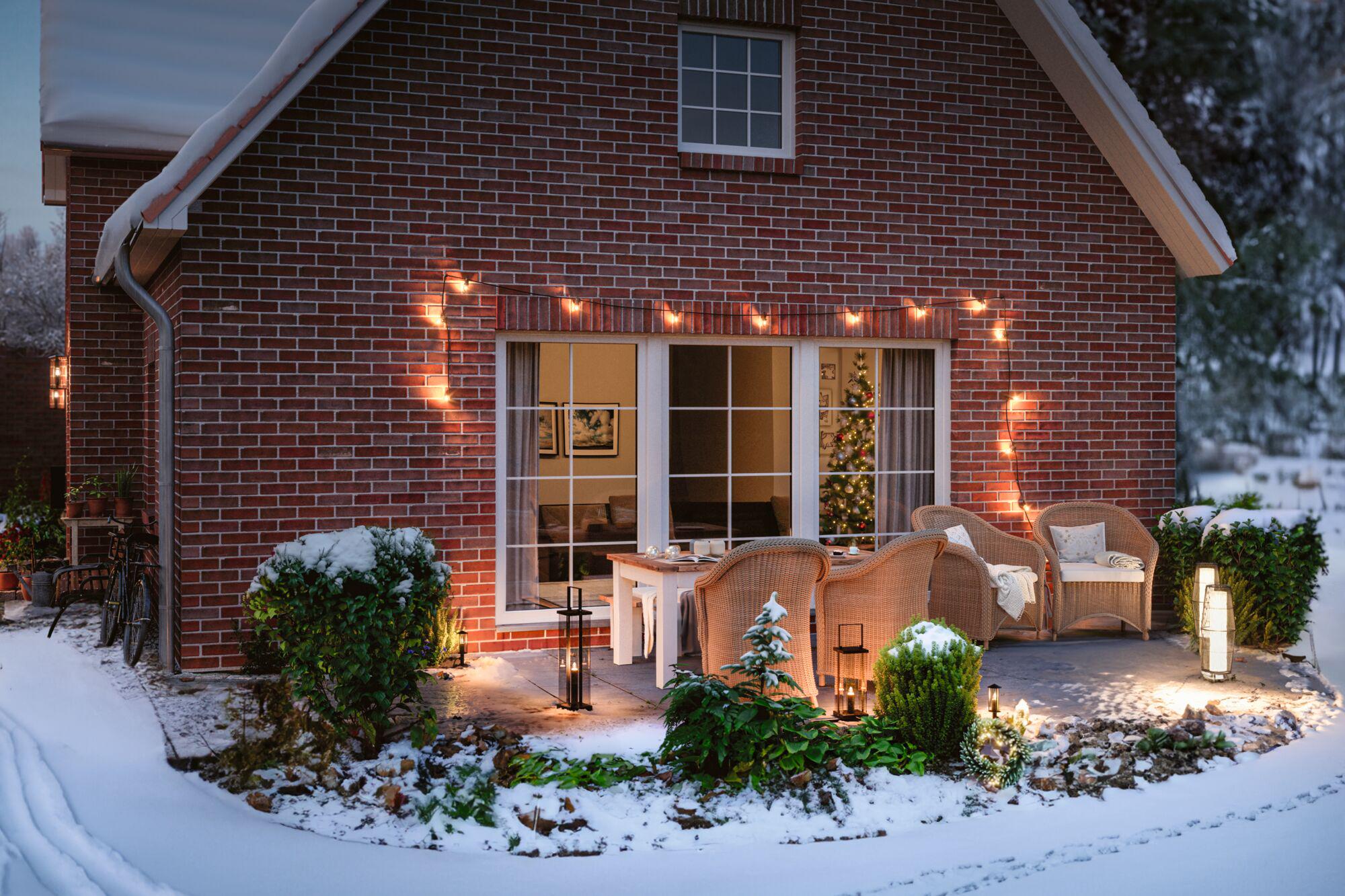 Gemütliche Terrasse mit Rattanmöbeln, Lichterkette und Laternen vor rotem Backsteinhaus im Schnee zur Weihnachtszeit