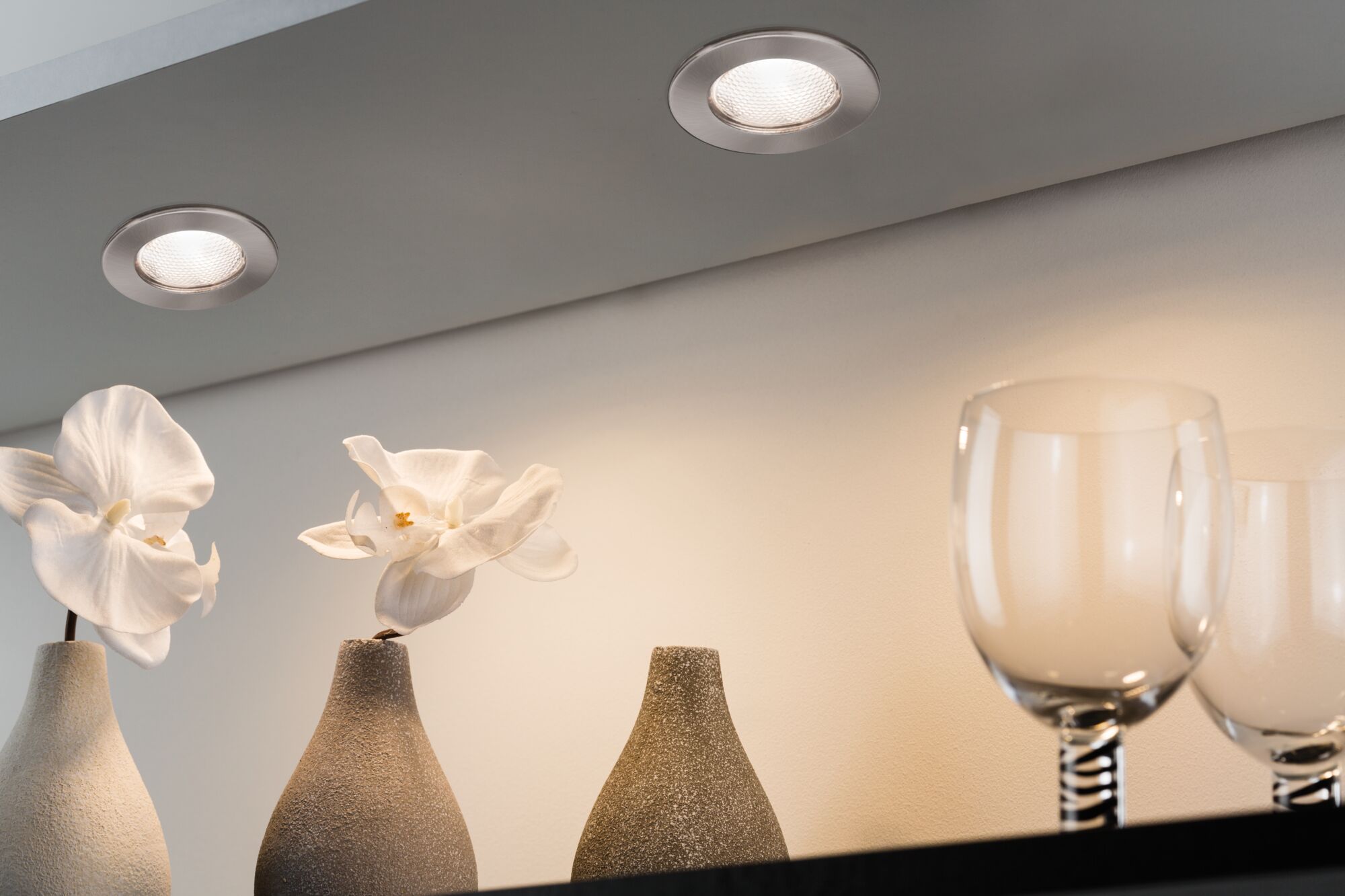 Two round brushed metal recessed lights illuminating white orchids in gray vases and glassware.