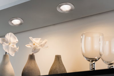 Two round brushed metal recessed lights illuminating white orchids in gray vases and glassware.