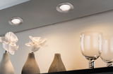 Two round brushed metal recessed lights illuminating white orchids in gray vases and glassware.