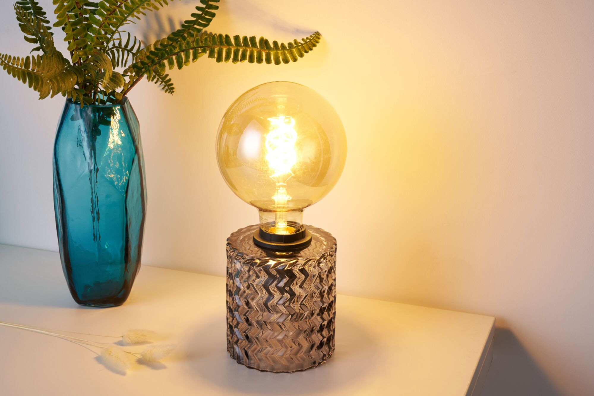 Table lamp with clear patterned glass base and warm LED bulb beside blue glass vase with fern leaves.