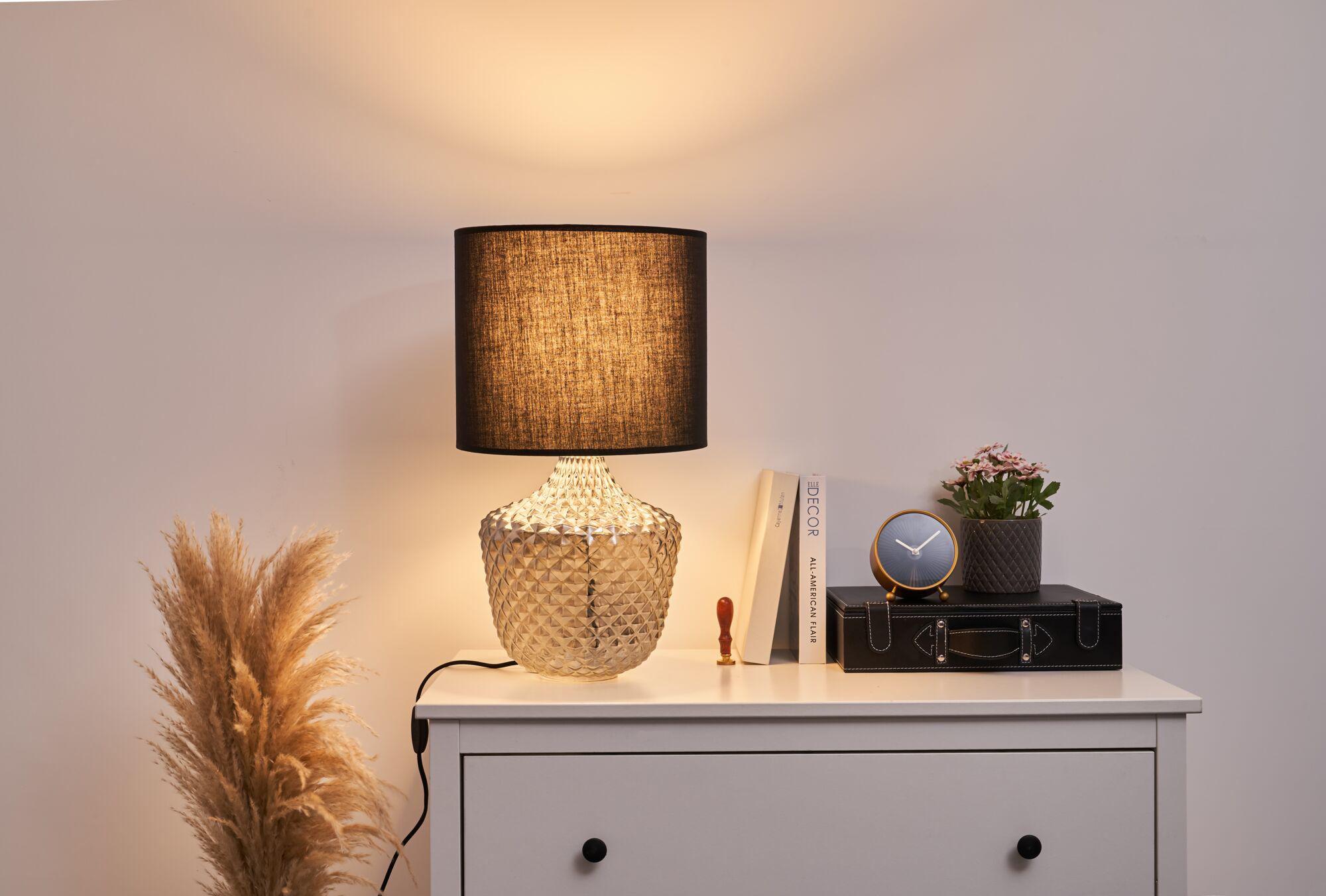 Table lamp with black fabric shade and textured glass base on white nightstand with decor items.