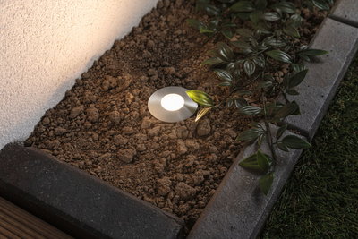 Spot encastré rond en acier inoxydable avec LED blanc chaud dans un parterre de jardin près d'une plante et d'un chemin en pierre.