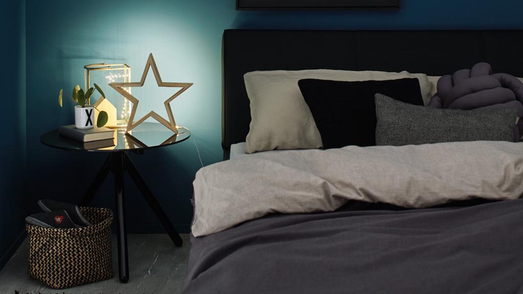 Gemütliches Schlafzimmer mit schwarzem Bett, grauer Bettwäsche und beleuchtetem Stern aus Holz auf Nachttisch.