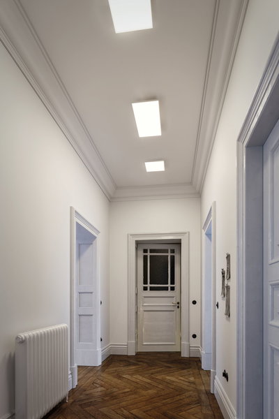 Couloir moderne avec murs blancs, sol en bois et luminaires rectangulaires au plafond pour éclairage intérieur