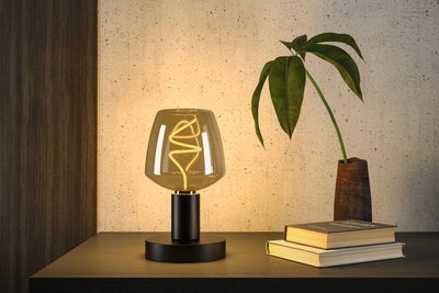 Moderne Tischlampe mit schwarzem Metallfuß und warmem Glaslicht neben Pflanze und Büchern auf Holzoberfläche